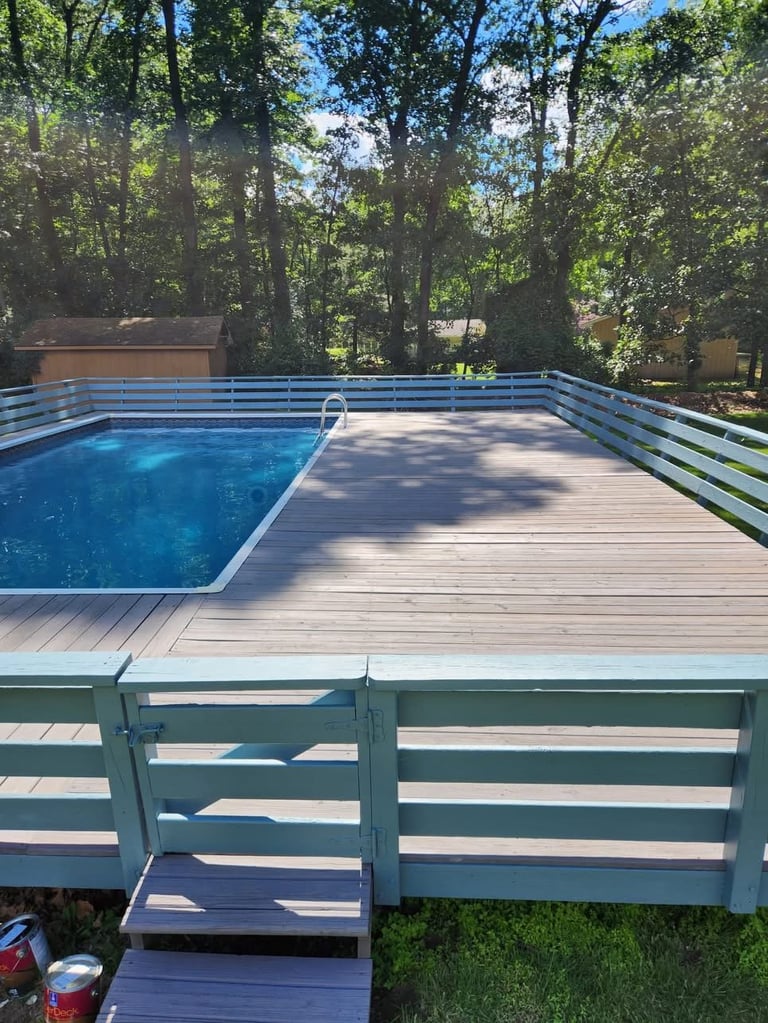 Blue above-ground swimming pool with gray wooden deck surrounded by tall trees by Central Ohio Deck Staining